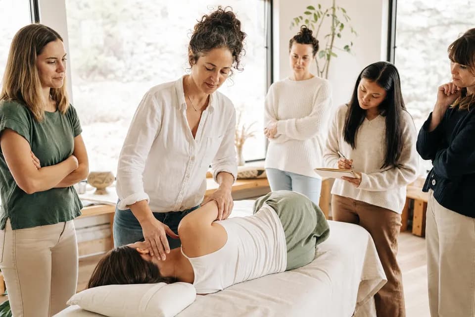 An instructor demonstrating a shoulder technique to bodywork students gathered around a treatment table